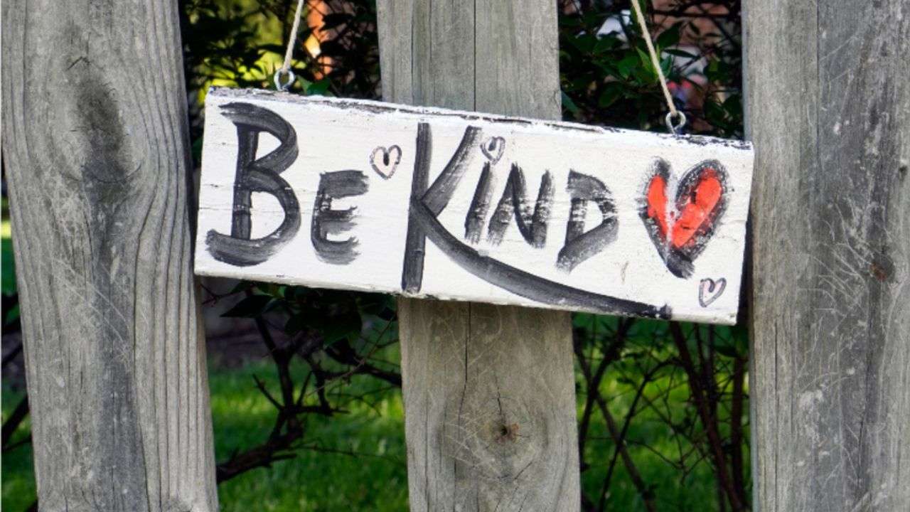 Be kind with a red heart painted on a sign which is hanging on a wooden fence